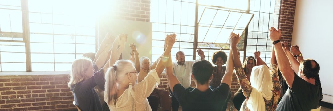 A diverse group of people holding hands in a circle, celebrating unity and teamwork in a sunlit room with large windows and brick walls. Diverse people sitting in a circle.