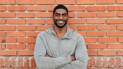 Obraz premium African American man in gray hoodie smiling against a brick wall. Casual African American man pose, arms crossed standing outdoor. Relaxed African American man wearing gray sport hoodie, smiling.