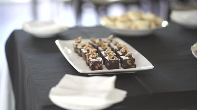Row of rich chocolate squares topped with walnut pieces sits neatly on a white plate over black linen, with other breakfast items softly blurred in the background, slow motion