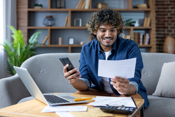 A happy man examines a bill while using his smartphone and laptop, working on his finances from home.