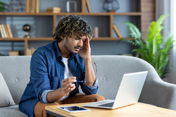 Obraz premium A young man, possibly experiencing a headache, sits near a laptop computer in a living room environment.