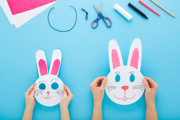 Mother and child hands together making white smiling rabbit paper masks on light blue table background. Pastel color. Point of view shot. Closeup. Creating easter decoration. Top down view.