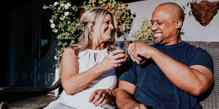 A couple enjoying drinks outdoors, smiling and toasting. Relaxed atmosphere with greenery. Happy couple, outdoor drinks, and smiles. Couple sitting together at night, diverse happy couple. - Powered by Adobe