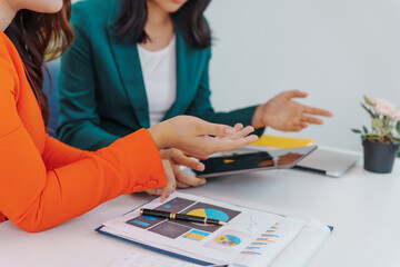 Two Asian businesswomen sit together at a desk, working professionally to solve problems. One...
