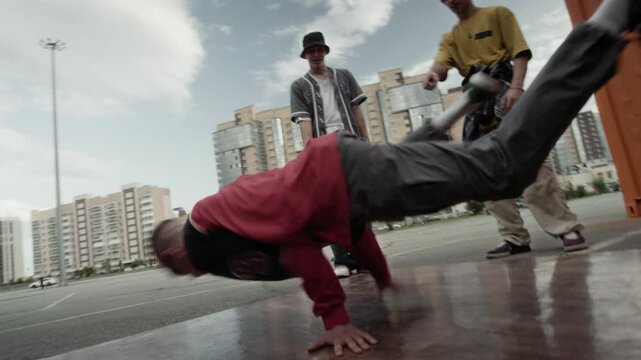 Handheld shot of talented Caucasian gen Z b-boy in street style clothes breaking outdoors in urban environment to hip hop music on boombox among friends cheering him up