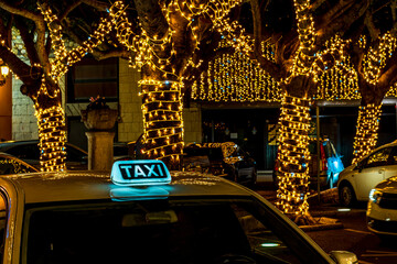  taxi with a lit-up TAXI sign on the roof. The taxi is parked in a city at holiday night, with...