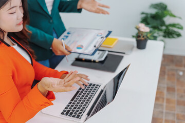 Two Asian businesswomen sit together at a desk, working professionally to solve problems. One...