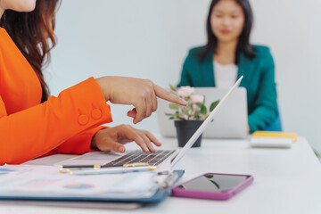 Two Asian businesswomen sit together at a desk, working professionally to solve problems. One...