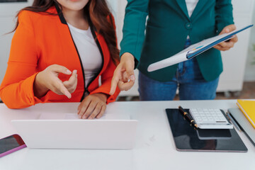 Two Asian businesswomen sit together at a desk, working professionally to solve problems. One...