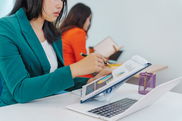 Two Asian businesswomen sit together at a desk, working professionally to solve problems. One...