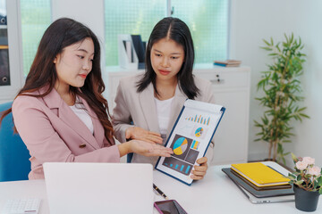 Two Asian businesswomen working together at a desk, focused and professional. They collaborate on...