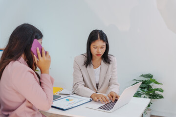 Two Asian businesswomen working together at a desk, focused and professional. They collaborate on...