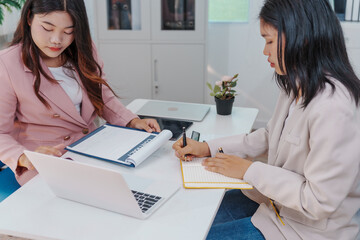 Two Asian businesswomen working together at a desk, focused and professional. They collaborate on...