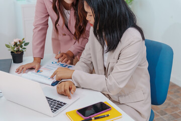 Two Asian businesswomen working together at a desk, focused and professional. They collaborate on...