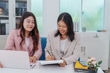 Two Asian businesswomen working together at a desk, focused and professional. They collaborate on...
