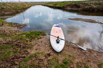 A serene paddleboarding scene unfolds by a tranquil waterway, surrounded by lush greens, inviting...
