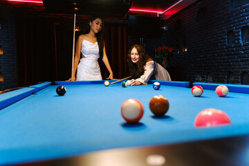 Fototapeta premium Two stylish women playing billiards in a modern, neon-lit bar lounge. One aims to strike the cue ball while the other observes confidently, capturing nightlife, friendship, and fun leisure activity.