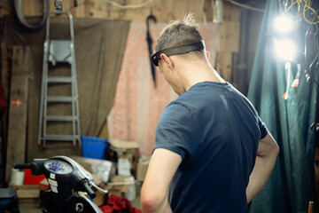 An Enthusiastic Mechanic Examining His Motorcycle in a Cluttered Garage Workshop with Tools and...