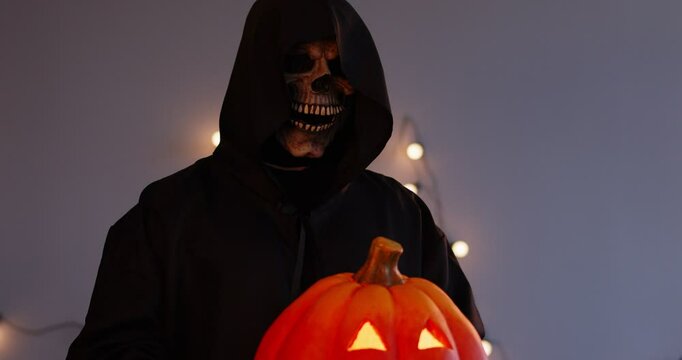 A man in a black suit and death mask holds an orange pumpkin. Halloween concept, scary pumpkin, halloween pumpkin head. Fantastic symbol of the angel of death. Scary mystical character