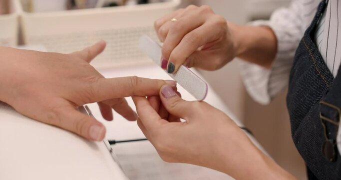 Close-up of Asian spa therapist woman filing senior customer&rsquo;s fingernails, hands moving precisely and smoothly across nail surface during manicure treatment inside cozy beauty salon environment.