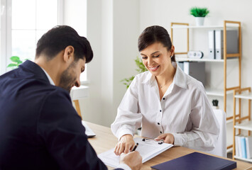 Fototapeta premium Young smiling businesswoman sitting at office desk, holding clipboard, showing where to put signature, while businessman signing contract, partner deal or employment agreement after successful meeting
