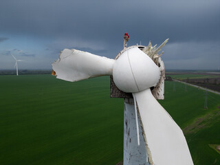 Close-up of a damaged wind turbine blade on a large green field with multiple turbines and cloudy...