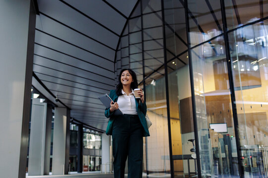 Businesswoman walking outside a modern office building holding a laptop and coffee