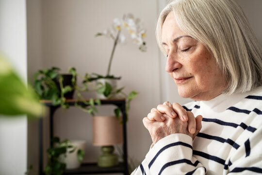 Elderly woman praying peacefully at home with eyes closed