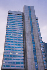 Obraz premium A sleek high rise building in Frankfurt, Germany, with curved edges, glass and concrete facade, vertical blue accents, and illuminated windows at dusk.
