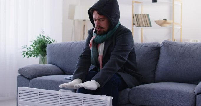 Freezing man wearing warm winter coat, scarf and gloves sitting on sofa near electric heater and trying to keep warm by rubbing and blowing into his hands, while shivering from cold. Heating problems.