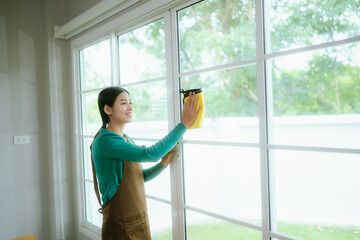 Smiling female cleaner in apron and gloves performs housework, dusting and disinfecting apartment...