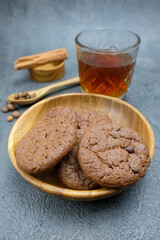 A freshly baked chocolate cookies served in a wooden bowl, accompanied by a glass of tea, cinnamon sticks, and coffee beans on a textured dark grey surface
