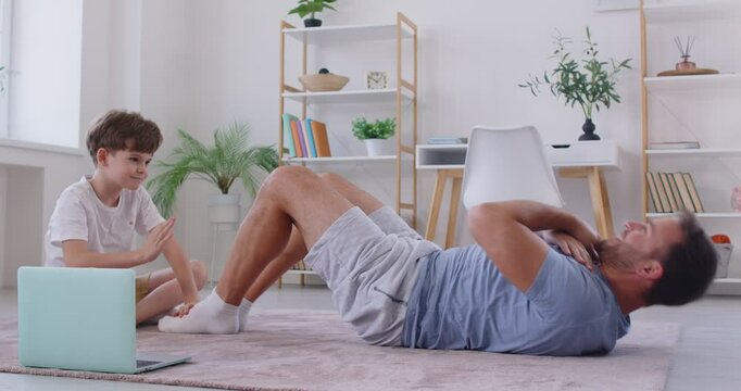 Sporty family, strong father practicing fitness doing abdominal sit ups, rock press exercise, working out wearing white sportswear, home interior, healthy lifestyle with little son giving high five