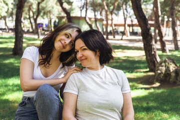 Fototapeta premium Two women sit closely together in a serene park setting, enjoying warm sunlight filtering through trees.
