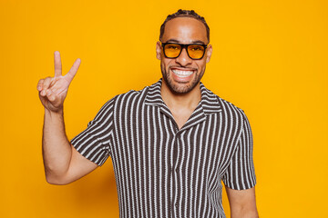 Naklejka premium Portrait of smiling young multiracial man wearing sunglasses showing victory sign while standing against yellow background at studio. 