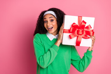 Excited young woman holding a gift box with a red bow, happy gesture on a pink background.