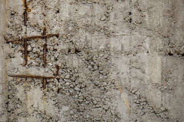 Rusted rebar grid protrudes from a unfinished, weathered concrete wall, forming an orange brown hued tic-tac-toe matrix in the wall's coarse monotone gray surface, texture with various small stones.