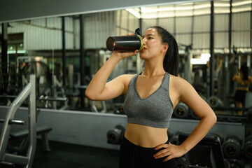 Confident young woman in sportswear enjoying a protein shake after a workout in a gym. Promotes...