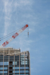 Tower crane stretches above construction of modern skyscraper with glass panels and scaffolding visible at top. Its red and white arm reaches into the clear blue sky, showcasing urban development.