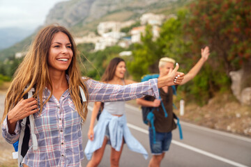 Cheerful interracial friends standing on road showing hitchhiking gestures. Tourism traveling