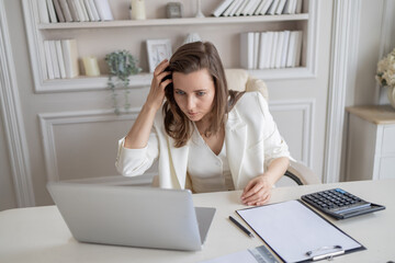 A woman in a white suit, a businesswoman, is sitting at a desk in an office, using a laptop screen as a mirror, preening herself. There is a laptop, a notebook, a pen and a calculator on the table.