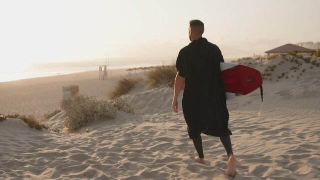 A surfer walks across the sand dunes towards the ocean at sunset, carrying his surfboard. He wears a black hooded poncho towel.