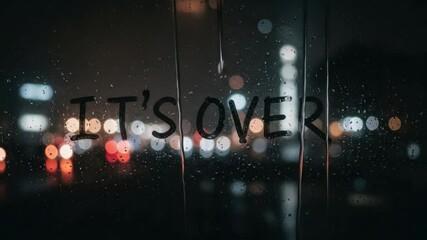 It's Over - Rainy Night Cityscape - The words "It's Over" are written on a rain-streaked window at night.  Out of focus city lights blur in the background, creating a melancholic atmosphere.
