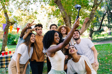 Group of cheerful young multiracial people taking a selfie with action camera mounted on a stick, having fun together at a party in a park