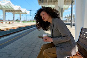 Young businesswoman with curly hair sitting on a bench at a train station, holding a coffee cup and...