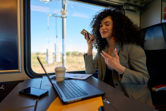 Businesswoman using laptop and talking on mobile phone while enjoying coffee during train journey, showcasing efficient remote work and travel productivity