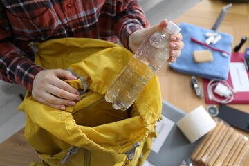 Survival kit. Woman putting bottle of water into backpack at table indoors, above view