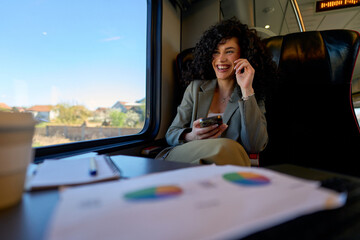 Businesswoman laughing while using her smartphone in a train seat, surrounded by business documents...