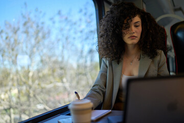Focused businesswoman working on a laptop and jotting down notes while enjoying a scenic train...