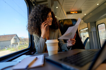 Businesswoman multitasking on a train, talking on her phone while reviewing documents and working on her laptop, enjoying a coffee during her commute